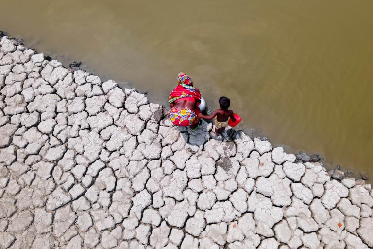 A woman in colorful clothing and a child sit on a cracked, dry riverbed, viewed from above.