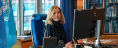 A person sitting at a desk, working on a computer in an office. A United Nations flag and bookshelf are visible in the background.