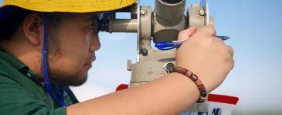 A person wearing a yellow hat looks through a surveying instrument while holding a pen and clipboard outdoors.