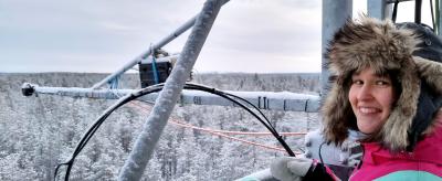 A person in a fur-lined hooded coat stands on a metal structure above a snowy forest, smiling at the camera.