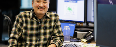 A man in a plaid shirt sits and smiles at a desk with multiple computer monitors and papers in an office setting.