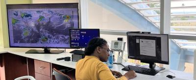 A person in a yellow sweater works at a desk with two monitors; one displays a weather map, the other shows a document. A large window and portable air conditioner are visible.