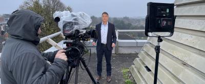 A man in a suit stands on a rooftop being filmed by a camera operator; professional lighting equipment is set up, and the weather appears overcast.