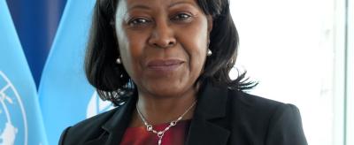A woman in professional attire stands in front of United Nations flags, looking at the camera.
