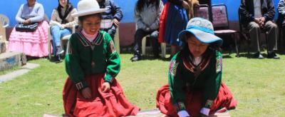 Two girls in traditional Andean clothing play a game with small objects on a woven mat outdoors, while people sit and watch in the background.