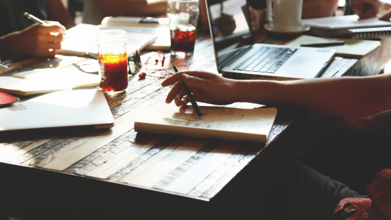 People sitting around a wooden table with laptops, notebooks, and drinks, engaged in discussion or work.