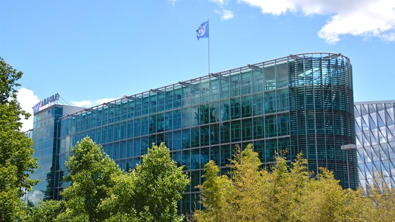 A modern glass office building with a blue company flag on the roof, partially obscured by trees under a bright sky with clouds.