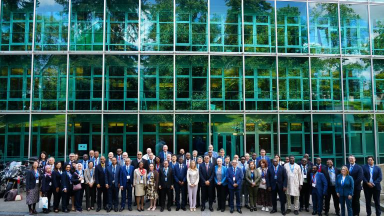A large group of formally dressed people poses for a photo in front of a modern glass building with green window frames.