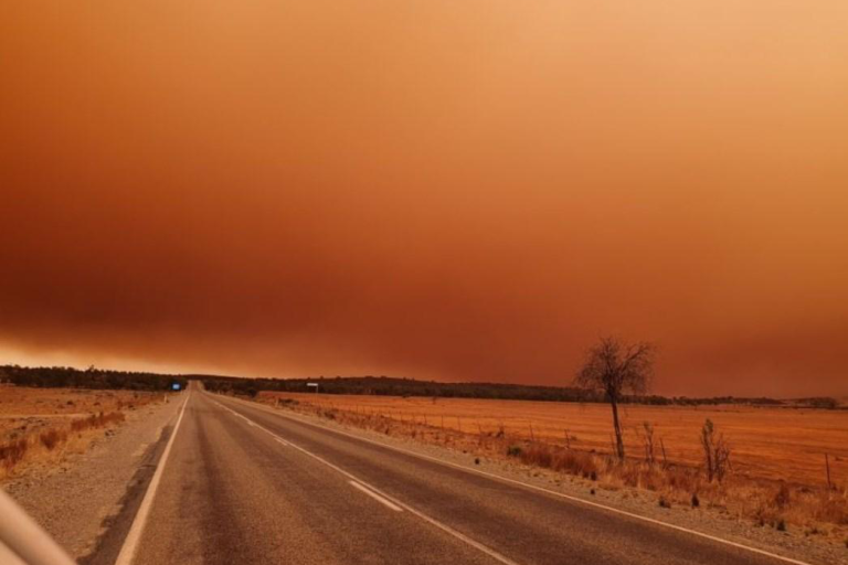 A car is driving down a road with a large orange dust storm in the distance.