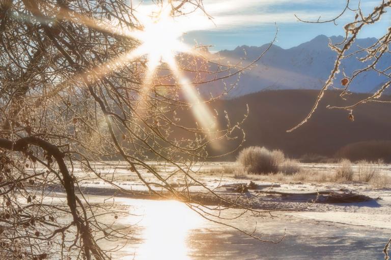 The sun is shining over a frozen river with mountains in the background.