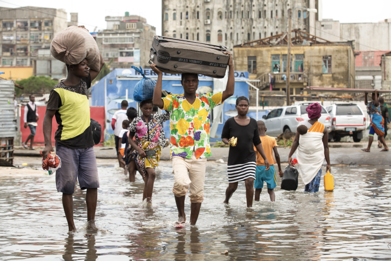People wade through a flooded street carrying goods on their heads and in their hands in an urban setting.
