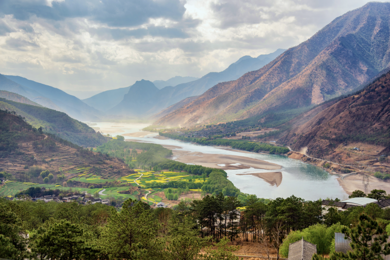 Scenic view of a winding river flowing through a valley with patchwork fields and mountain ranges under a cloudy sky.