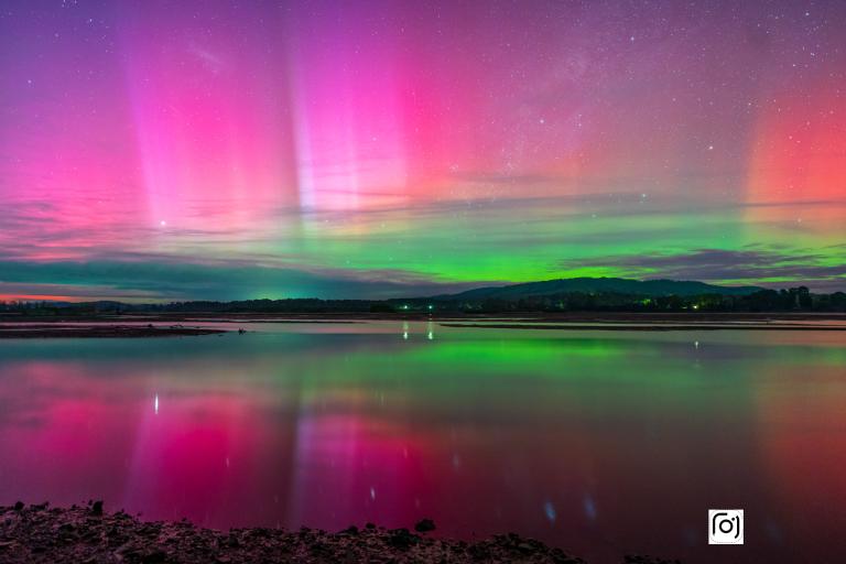 Vibrant aurora borealis reflected over a tranquil lake, with pink and green hues in the sky and a serene landscape in the background.