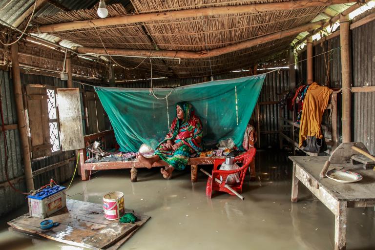 A person sits on a bed under a mosquito net inside a flooded wooden and metal hut. Household items are scattered, and water covers the floor.