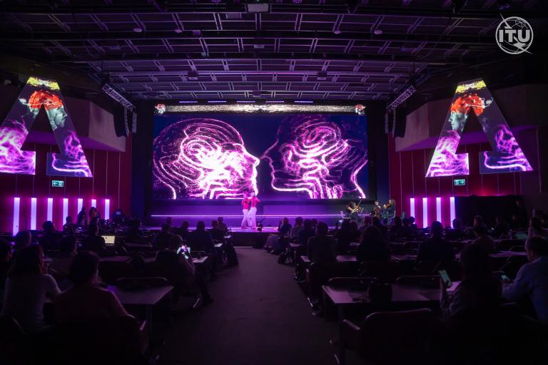 A speaker presents on a stage with a large screen displaying neon graphics of two human heads in profile. The audience is seated and attentive in a dimly lit auditorium.