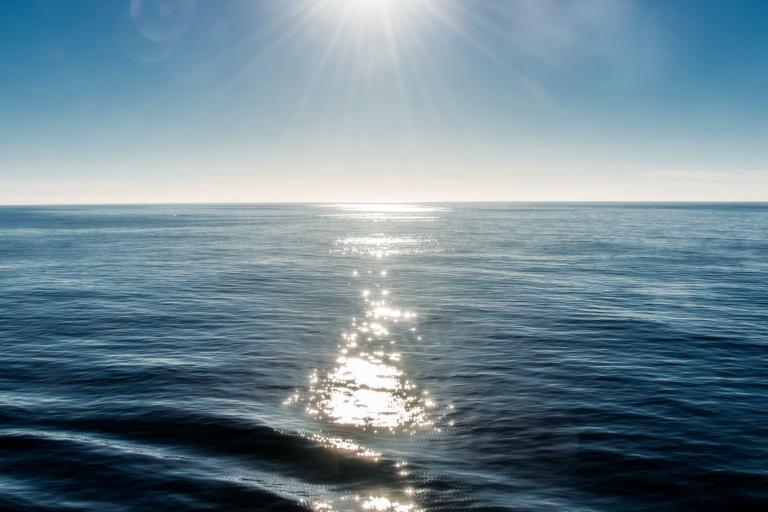 The sun shines brightly over a vast, calm ocean, creating a path of sparkling light on the water's surface.
