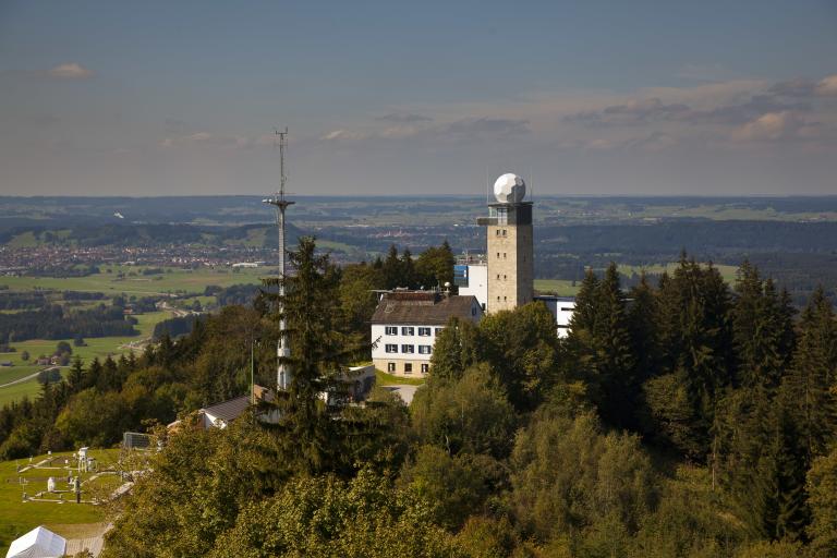 Aerial view of a hilly landscape with a meteorological tower, two buildings, forested areas, and distant fields on a clear day.
