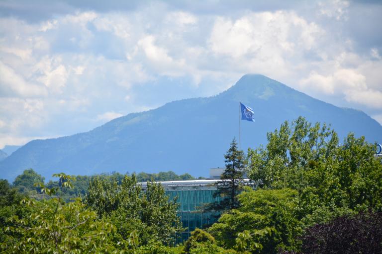 A glass building is surrounded by greenery with a flag flying in front of it. In the background, there is a mountain range under a partly cloudy sky.