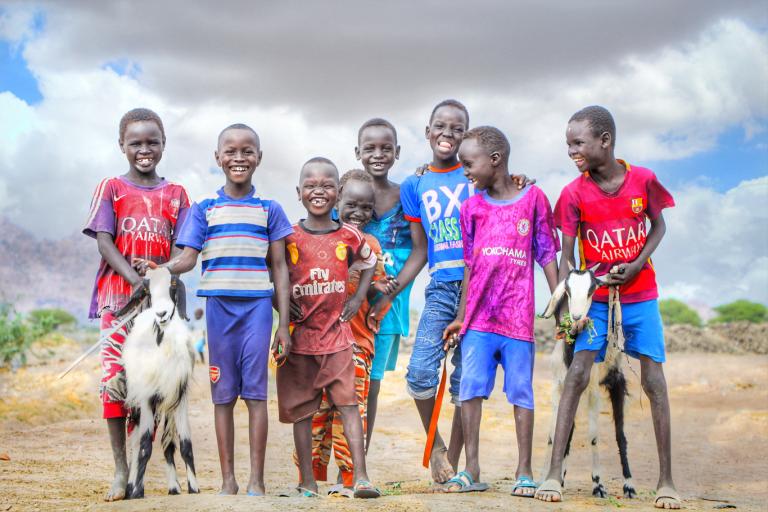 A group of seven children stands outdoors on a dirt path, smiling at the camera. Two of the children are holding goats. The sky is partly cloudy in the background.
