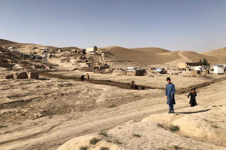 A man and a child walk on a dirt path in a rural, arid landscape with simple housing structures and people working in the distance.