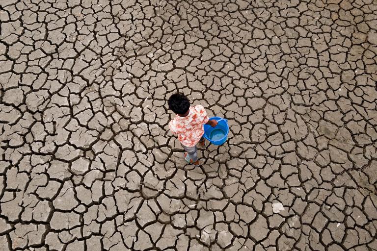 A person carrying a blue bucket walks on cracked, dry earth, suggesting a drought or water scarcity.