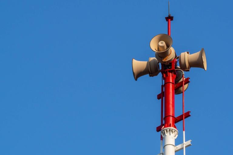 Four loudspeakers mounted on a red and white pole against a clear blue sky.