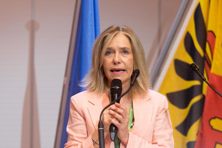 A woman in a light pink blazer speaks into a microphone at a podium, with blue and yellow flags in the background.