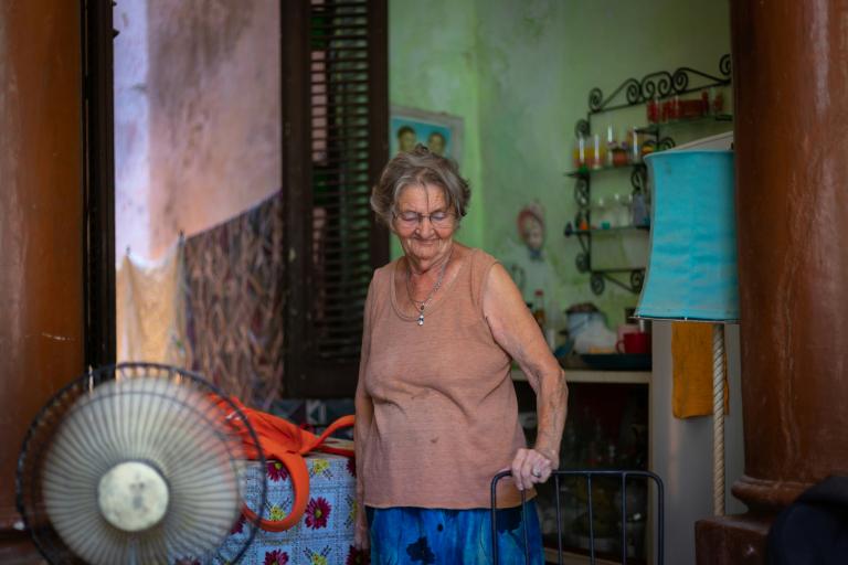 An elderly woman stands in a room with a fan, colorful cloth, and household items. She wears a sleeveless top and glasses, looking down with a gentle smile.