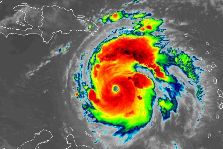 Satellite image of a large, well-defined hurricane with a visible eye, near the Caribbean islands, displaying intense colors indicating varying levels of precipitation and storm intensity.