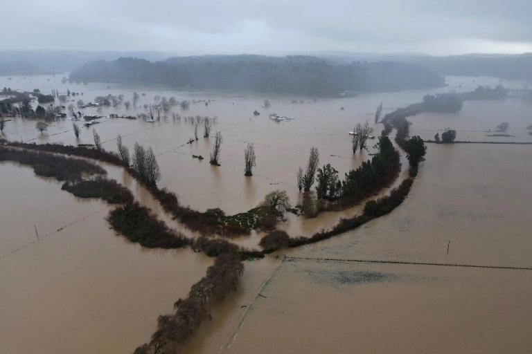 An aerial view of extensive flooding in a rural area of Chile, where fields are submerged under muddy water, and trees and structures are partially inundated.