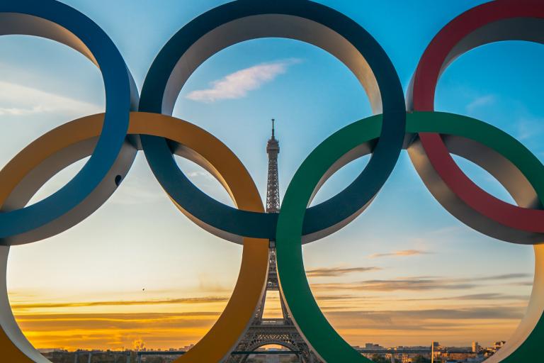 The Eiffel Tower is framed through the Olympic rings in Paris at sunset, with a colorful sky in the background.