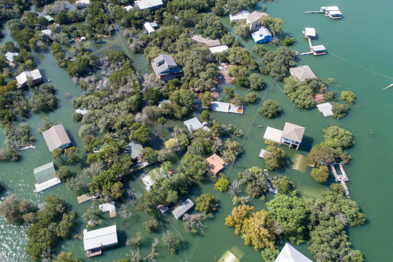 Aerial view of a flooded community with partially submerged houses and trees, surrounded by rising water levels. Some structures are completely surrounded by water.