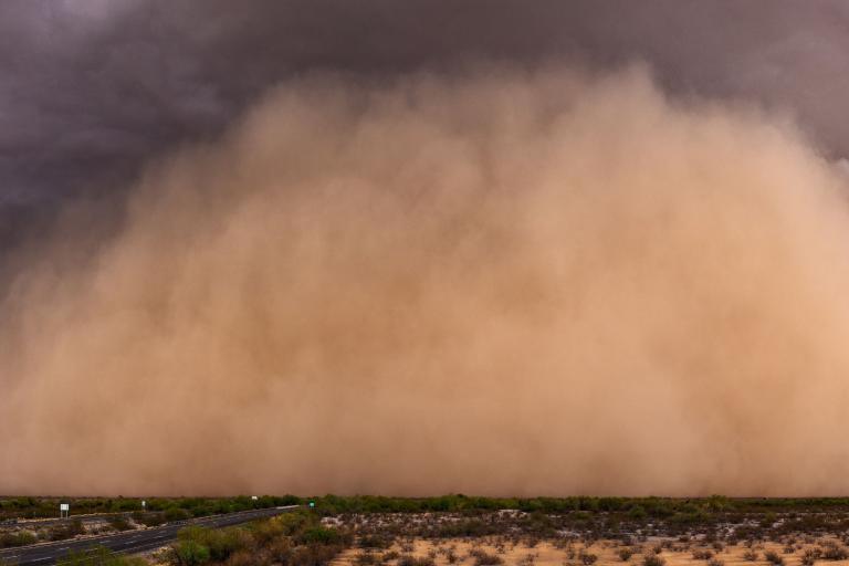 A large dust storm sweeps across a barren, desert landscape with a road on the left side of the image. The sky is dark and ominous.