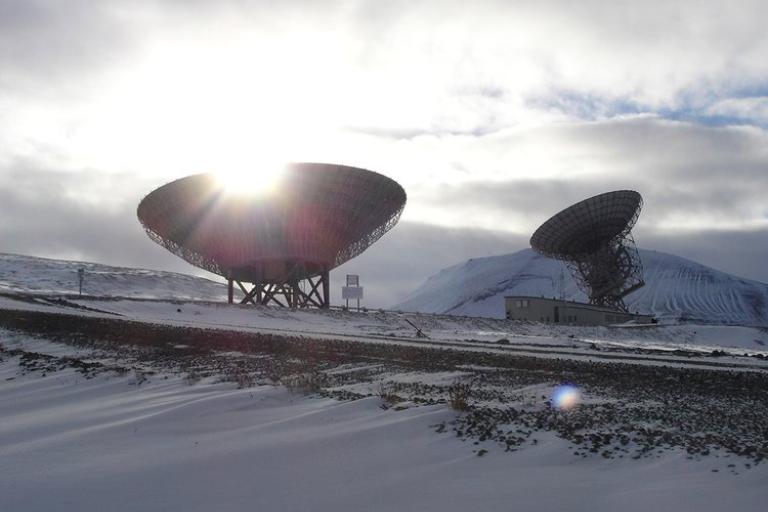 Two large satellite dishes are situated in a snowy landscape with mountains in the background. The sun is partially visible behind one of the dishes, casting light through its structure.