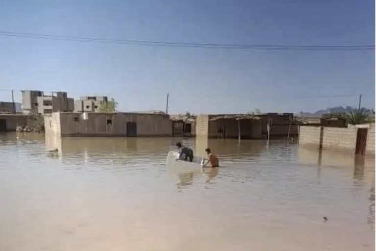 Three people in a small boat navigate through a flooded area with partially submerged buildings under a clear blue sky.