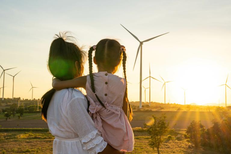 A woman with a young girl in her arms stands outdoors, looking at wind turbines during sunset.