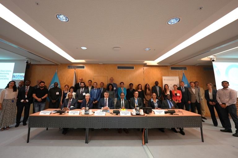 A group of individuals stand behind a conference table with microphones and nameplates in a formal meeting room. Participants are dressed in business attire.