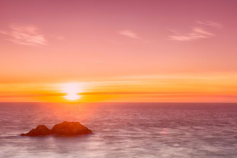 A serene sunset over a calm ocean with two small rocks in the foreground, under a pink and orange sky.