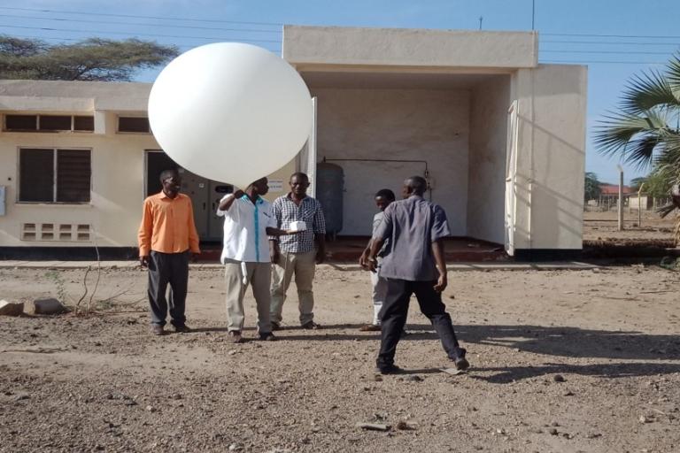 A group of people prepares to launch a large white weather balloon outside a small building on a sunny day.