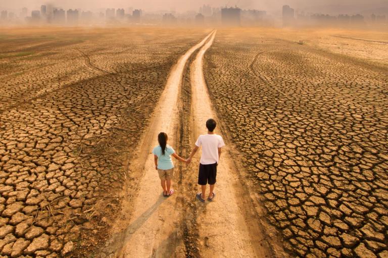 Two people stand on a dry, cracked path leading toward a distant city skyline under a hazy sky.