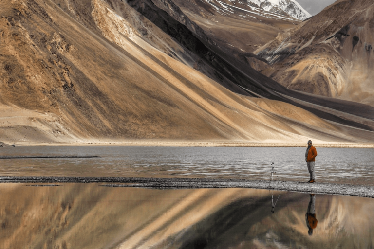 A person stands near a tripod by a reflective mountain lake with snow-covered peaks in the background.