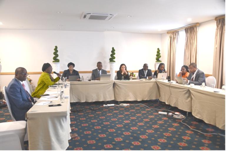 A group of people sit around a U-shaped conference table in a meeting room, each with laptops or documents, engaged in discussion.
