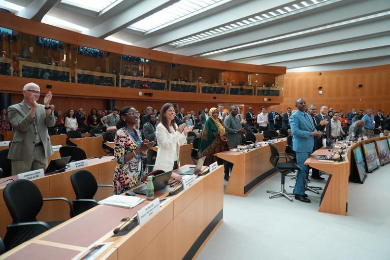 A group of people in business attire stands and applauds inside a modern conference room with wooden walls and large windows.