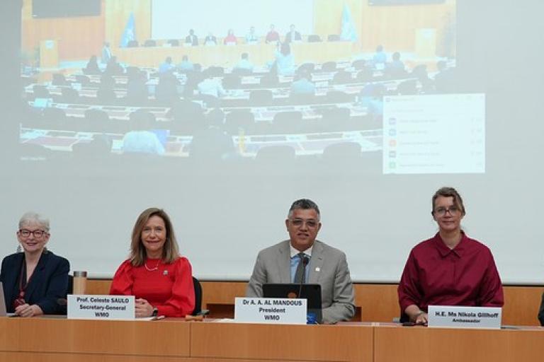 Six people sit at a conference table with nameplates, facing the camera, with a large screen showing an audience and meeting behind them.