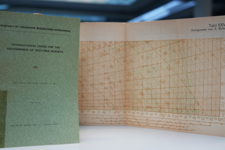 A green booklet titled "International Codes for the Transmission of Weather Reports" stands next to an open chart with grid lines and numerical data.