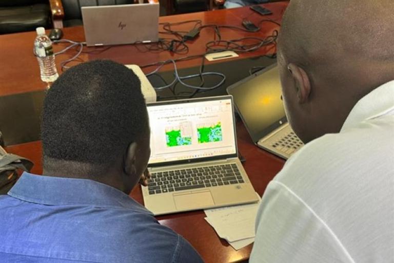 Two people look at weather or map data displayed on a laptop screen in an office meeting setting, with laptops and papers on the table.