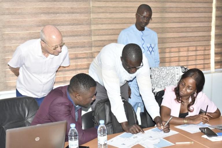 Five people are gathered around a table with papers, a laptop, and water bottles, engaged in a discussion and reviewing documents in an office setting.