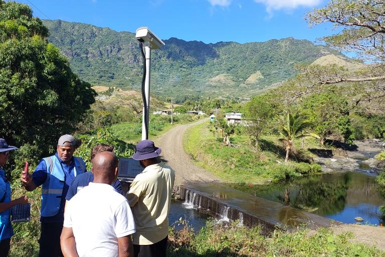 Four people stand near a small river and road, next to a tall monitoring device, with mountains and trees in the background on a sunny day.