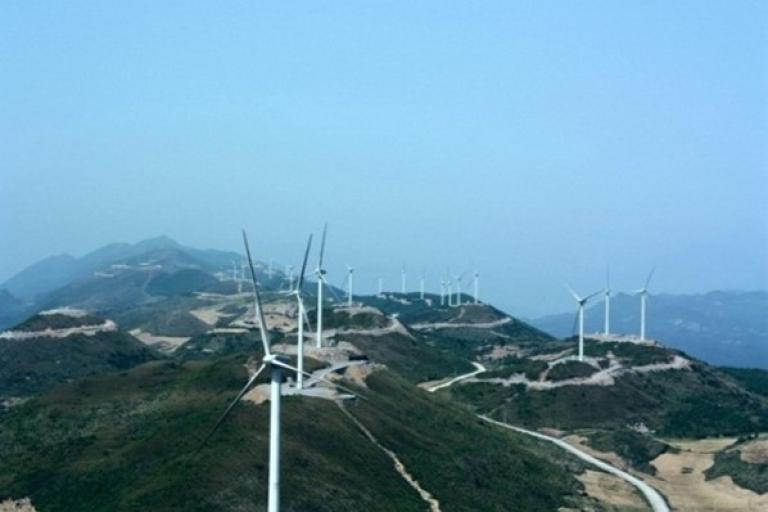 A series of wind turbines are installed along a hilly landscape, with winding roads running through the area under a clear sky.