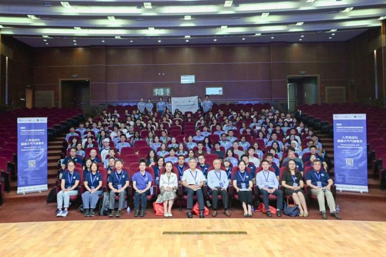 A large group of people pose for a photo in an auditorium, seated in rows of red chairs, with banners and a wooden stage visible in the foreground.